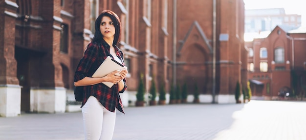 Young pensive woman looking away while holding digital tablet and standing at square in city