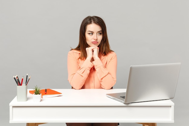 Young pensive woman in pastel casual clothes put hands prop up on chin, sit work at desk with pc laptop isolated on gray background. Achievement business career lifestyle concept. Mock up copy space.