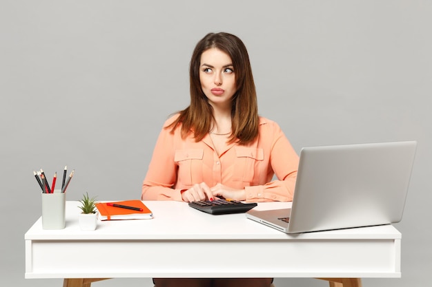 파스텔 캐주얼 옷을 입은 젊은 수심 있는 여성은 계산기를 사용하여 회색 배경에 격리된 PC 노트북을 들고 흰색 책상에 앉아 일합니다. 성취 비즈니스 경력 라이프 스타일 개념입니다. 복사 공간을 비웃습니다.