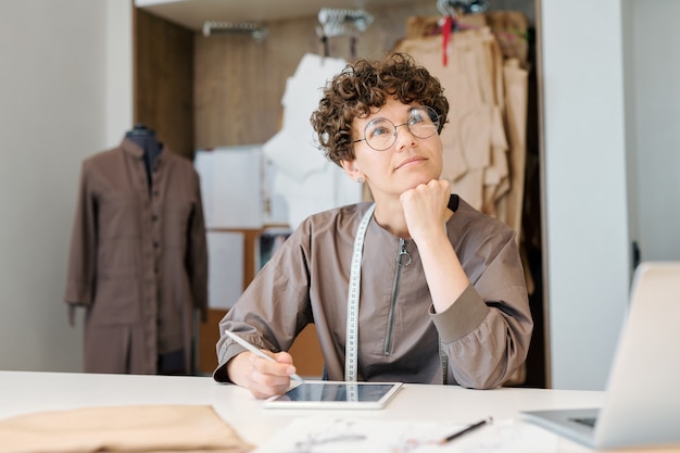 Young pensive woman thinking of new ideas while working over fashion models by desk in workshop