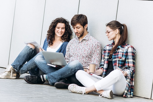 Young people are using different gadgets and smiling, sitting near the wall. Students studying using laptop computer. Education social media concept.