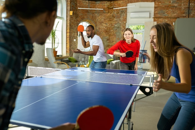 Young people playing table tennis in workplace, having fun. Friends in casual clothes play ping pong together at sunny day. Concept of leisure activity, sport, friendship, teambuilding, teamwork.