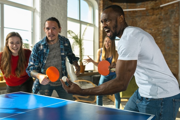 Молодые люди играют в настольный теннис на рабочем месте, веселятся Друзья в повседневной одежде играют