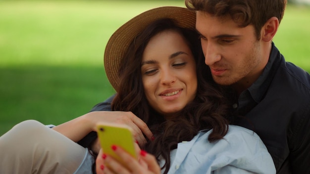 Young people scrolling phone in summer park Portrait of surprised guy pointing with finger on mobile phone screen outdoors Closeup attractive couple having rest with smartphone on picnic