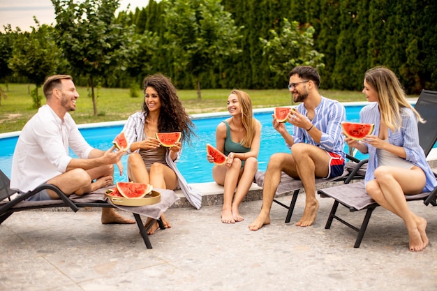 Young people sitting by the swimming pool and eating watermelon in the house backyard