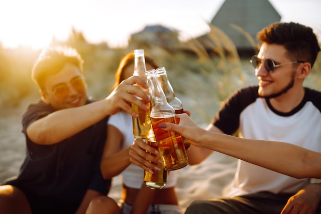 Молодые люди сидят вместе на пляже и пьют пиво Группа друзей приветствует пиво на пляже