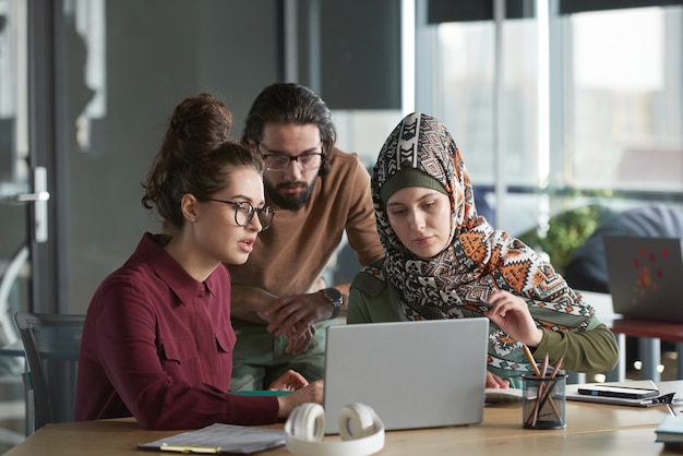 Young people together with muslim woman looking at monitor of laptop and working over online presentation at office