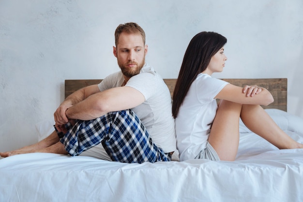 Young people wearing pajamas sitting on a bed with hands over their knees and looking serious after a conflict