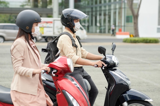 Молодые люди в защитных шлемах и медицинских масках едут на работу на скутерах.