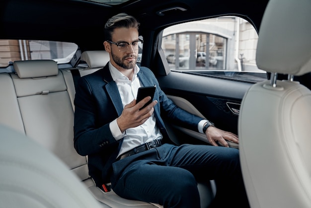 Young perfectionist. Handsome young man in full suit looking at his smart phone while sitting in the car