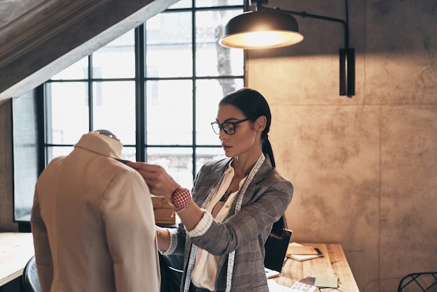 Young perfectionist. Serious young woman in eyewear adjusting a collar of the jacket on mannequin while standing in her workshop