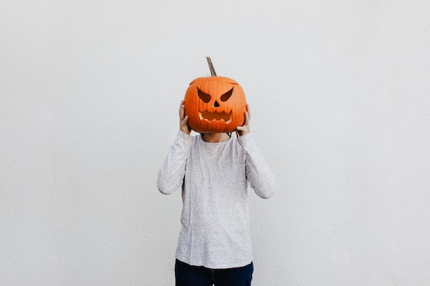 Young person holding a carved pumpkin head as his or her head\
on white wall background. halloween season mood.