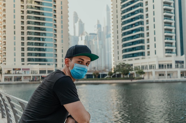 Young person in medical mask standing outdoor and looking at camera with urban city background