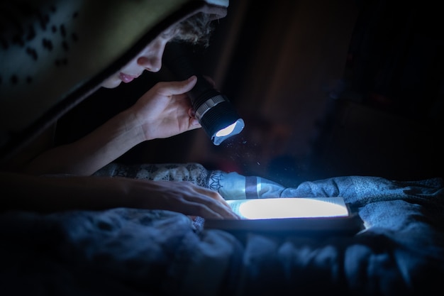 Young person with a light torch reading books in bed at night in bedroom