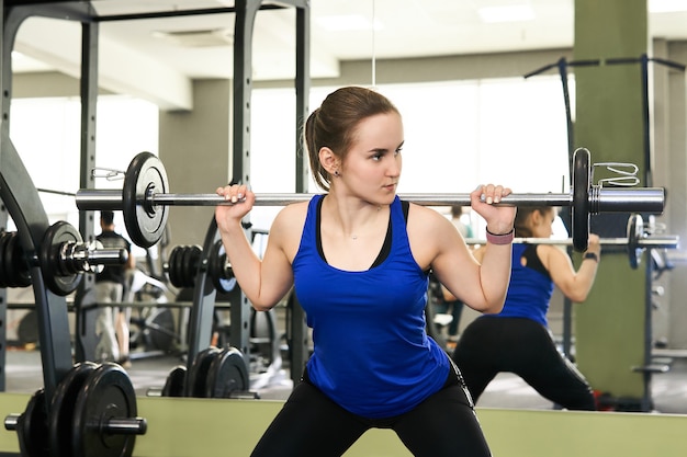 Young petite woman performs the exercise squat with barbell in the gym reflecting in the mirror