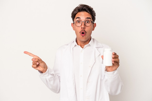 Молодой фармацевт смешанной расы мужчина держит таблетки, изолированные на белом фоне, указывая в сторону