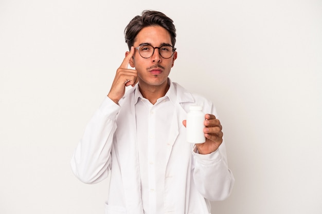 Young pharmacist mixed race man holding pills isolated on white background pointing temple with finger, thinking, focused on a task.