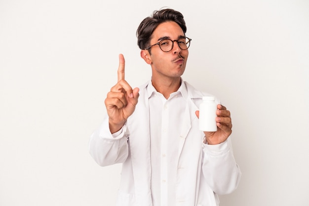 Young pharmacist mixed race man holding pills isolated on white background showing number one with finger.