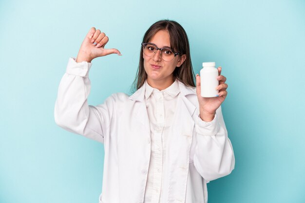 Young pharmacist woman holding pills isolated on blue background feels proud and self confident, example to follow.