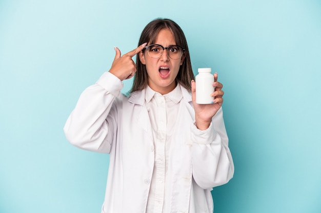 Young pharmacist woman holding pills isolated on blue background showing a disappointment gesture with forefinger.