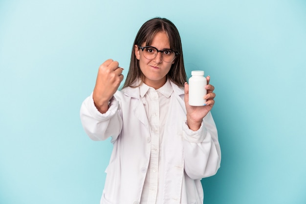 Young pharmacist woman holding pills isolated on blue background showing fist to camera, aggressive facial expression.