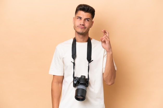 Photo young photographer caucasian man isolated on beige background with fingers crossing and wishing the best