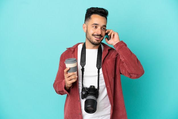 Young photographer caucasian man isolated on blue background holding coffee to take away and a mobile