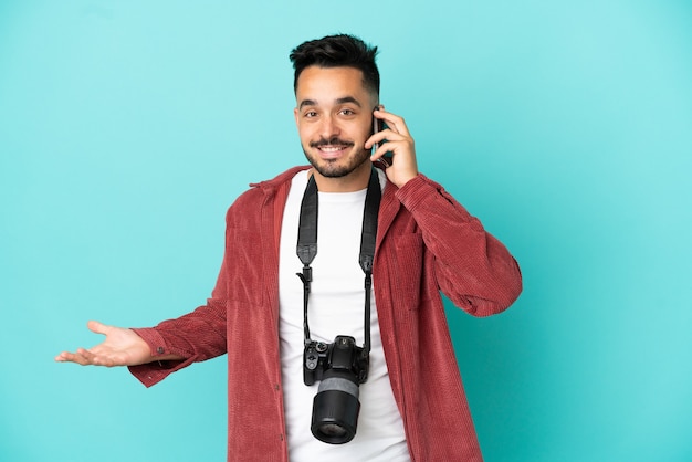 Young photographer caucasian man isolated on blue background keeping a conversation with the mobile phone with someone