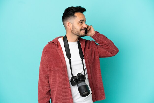 Young photographer caucasian man isolated on blue background keeping a conversation with the mobile phone