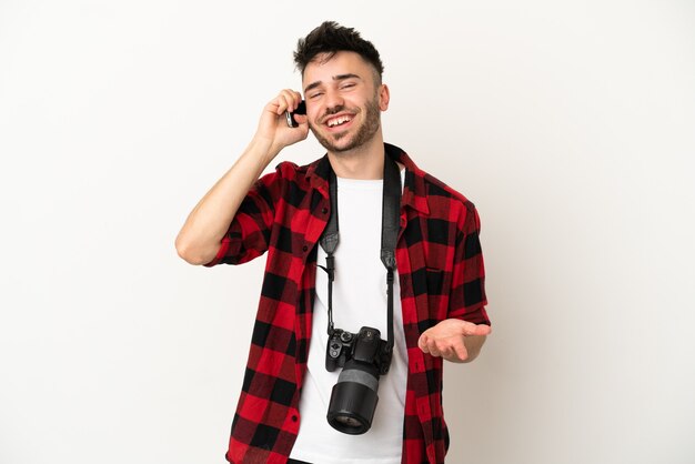 Giovane fotografo caucasico uomo isolato su sfondo bianco che tiene una conversazione con il telefono cellulare con qualcuno