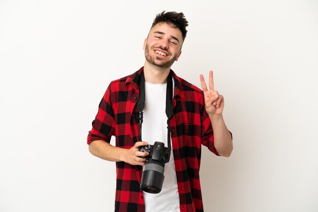 Uomo caucasico del giovane fotografo isolato su fondo bianco che sorride e che mostra il segno di vittoria