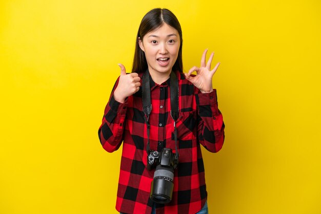 Young photographer chinese woman isolated on yellow background showing ok sign and thumb up gesture