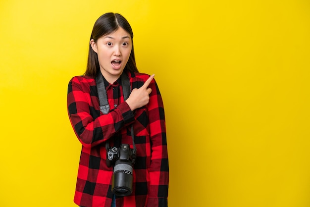 노란색 배경에 고립된 젊은 사진작가 중국 여성은 놀라고 측면을 가리키고 있습니다.