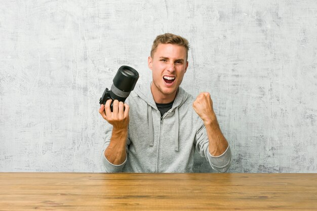 Young photographer holding a camera on a table cheering carefree and excited