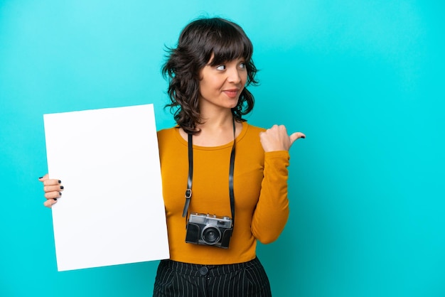 空のプラカードを保持し、側を指している青い背景に分離された若い写真家ラテン女性