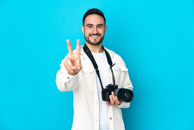 Young photographer man isolated on blue background smiling and showing victory sign