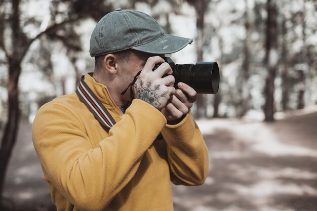 Молодой фотограф мужчина в шляпе работает, фотографируя природу в лесу, держа и используя свою камеру осенью или зимой