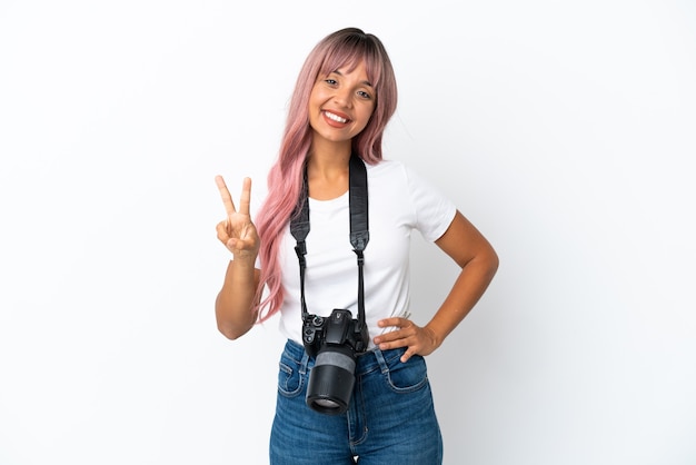 Молодой фотограф смешанной расы женщина с розовыми волосами на белом фоне улыбается и показывает знак победы