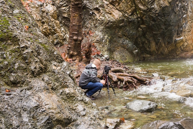 Foto giovane fotografo con lo zaino che fa le foto della cascata e delle rocce con la macchina fotografica.