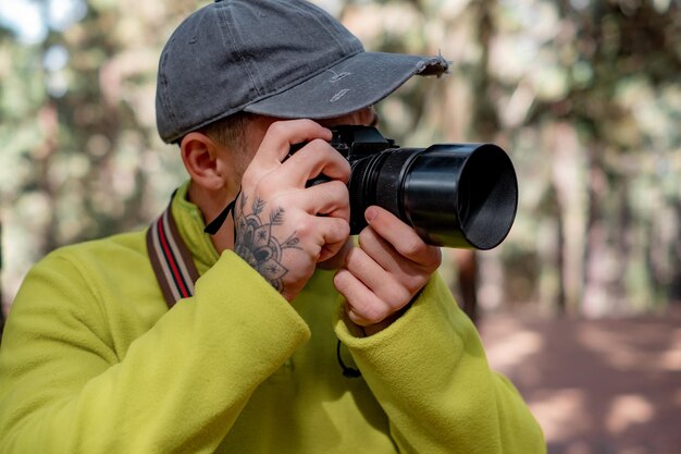 Молодой фотограф в шляпе работает, фотографируя природу в лесу, держа и используя камеру осенью или зимой