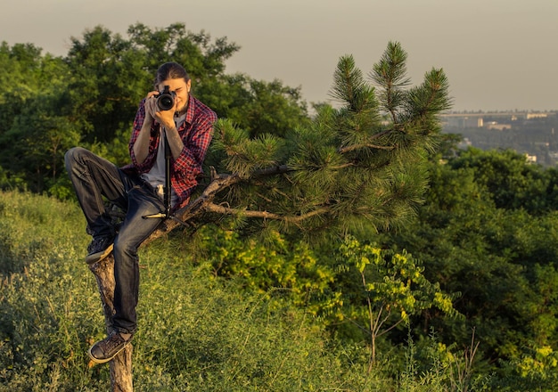 Young photographer with professional digital camera sitting on the tree and taking shot against park outdoors and city skylines