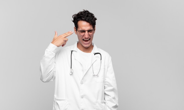 Young physician looking unhappy and stressed, suicide gesture making gun sign with hand, pointing to head