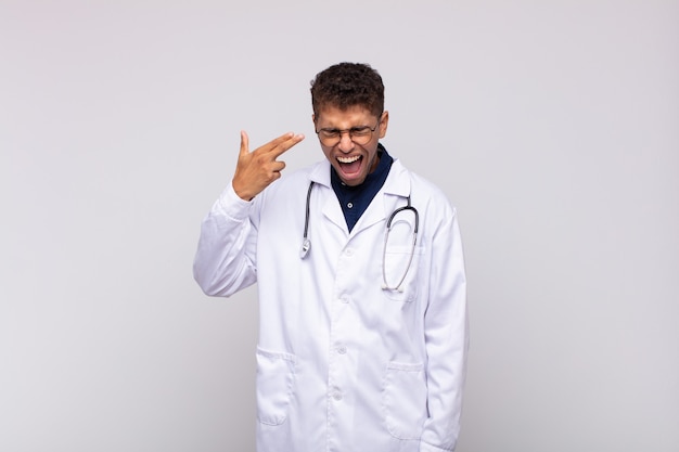 Young physician man looking unhappy and stressed, suicide gesture making gun sign with hand, pointing to head