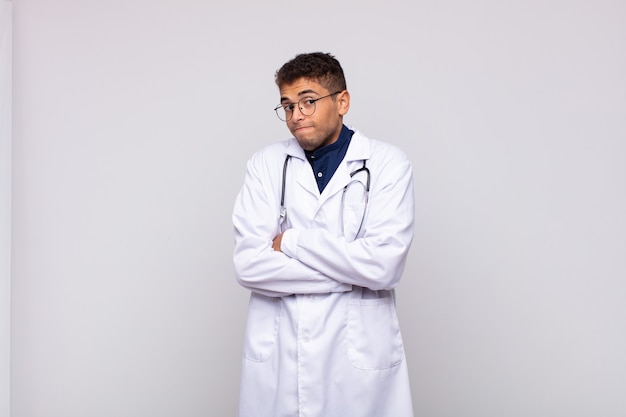 Young physician man shrugging, feeling confused and uncertain, doubting with arms crossed and puzzled look