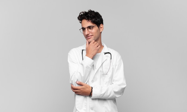 Young physician smiling with a happy, confident expression with hand on chin, wondering and looking to the side