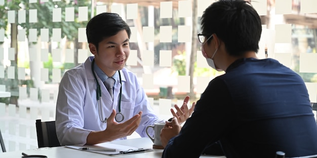 현대 의사를 통해 의사의 작업 책상에 함께 앉아있는 동안 바이러스와 전염병에 대해 환자에게 이야기 / 설명하는 젊은 의사