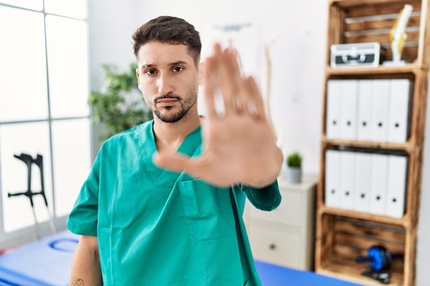 Young physiotherapist man working at pain recovery clinic doing stop sing with palm of the hand. warning expression with negative and serious gesture on the face.