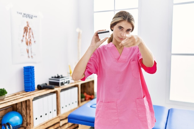 Молодая женщина-физиотерапевт, работающая в клинике восстановления боли, держа кредитную карту, указывая пальцем на камеру и на вас, уверенный жест выглядит серьезным