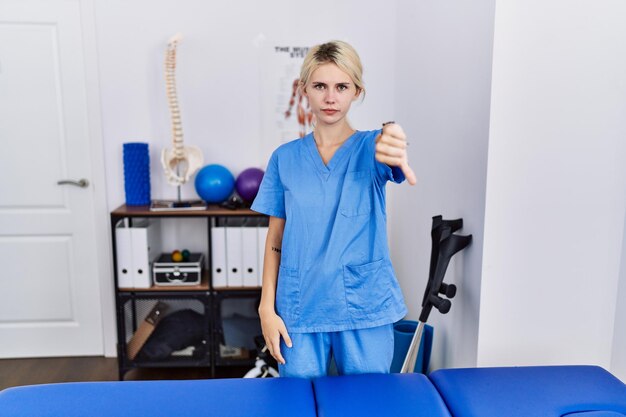 Young physiotherapist woman working at pain recovery clinic looking unhappy and angry showing rejection and negative with thumbs down gesture. bad expression.