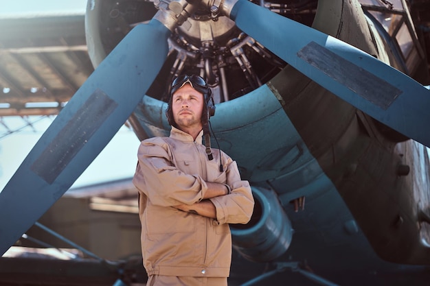 Photo young pilot is thoughtfully looks away. he is standing near small blue retro jet, just under the propeller.